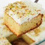 A square slice of pistachio pudding cake with light green frosting and chopped pistachios on top is being lifted from a baking dish, revealing a soft, moist texture inside.