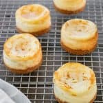 Five mini pumpkin swirl cheesecakes with golden-brown tops and crumbly crusts cool on a wire rack, with a light-colored cloth partially visible in the lower left corner.