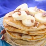 A stack of banana nut pancakes topped with banana slices and chopped pecans sits on a blue plate, with a purple cloth in the background.