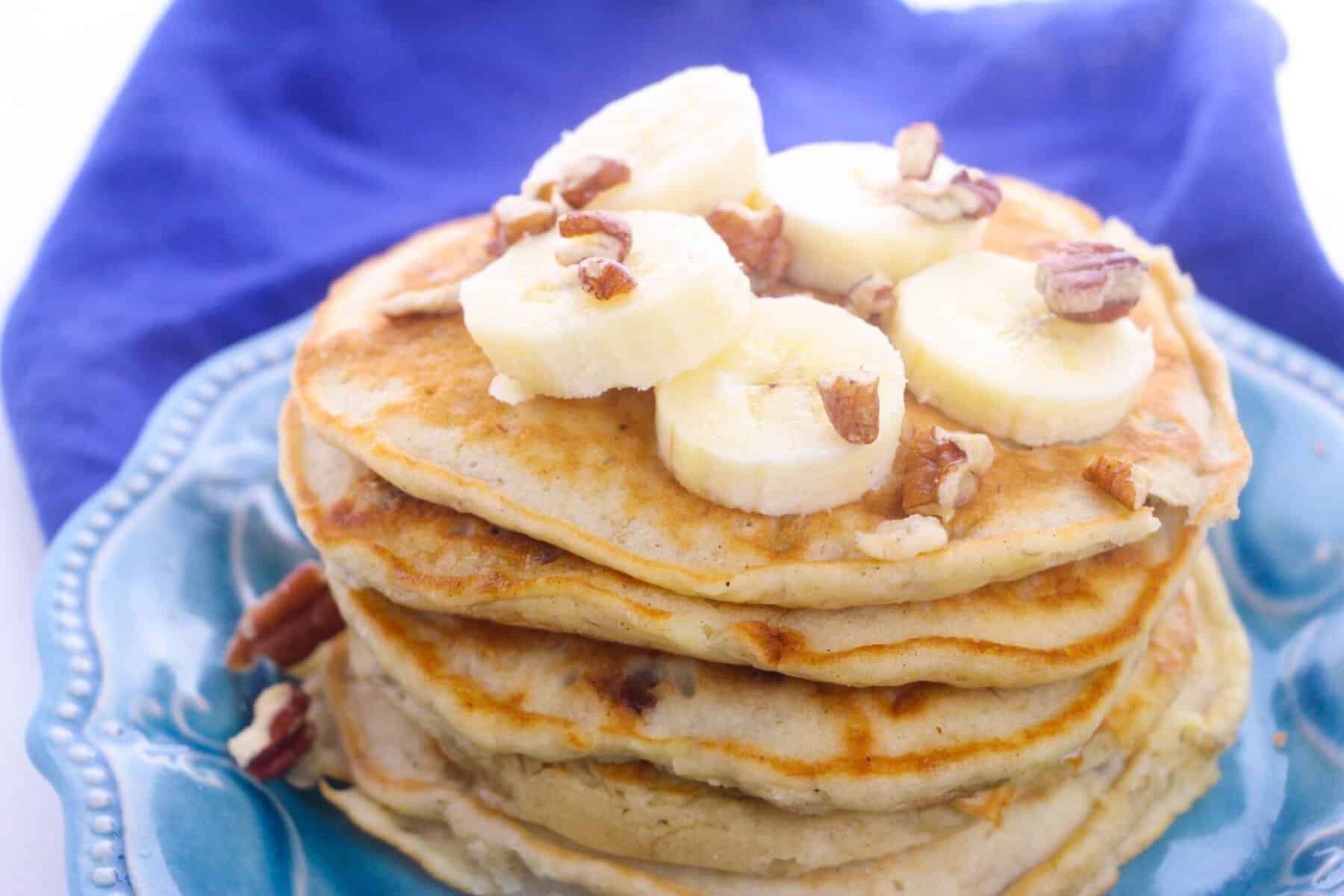 A stack of banana nut pancakes topped with banana slices and chopped pecans sits on a blue plate, with a purple cloth in the background.