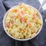 A white bowl filled with creamy macaroni salad, featuring diced red and green vegetables, is placed on a folded dark gray cloth with a spoon resting in the bowl.