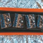 Four rectangular hocus pocus brownies decorated with blue icing and candy eyeballs sit on a black tray with an orange border, placed on a tablecloth patterned with black spiderwebs.