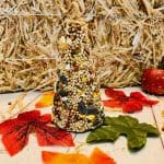 An ice cream cone bird feeder covered in seeds sits on a wooden surface, surrounded by colorful artificial autumn leaves, a straw bale, and small decorative gourds in the background.