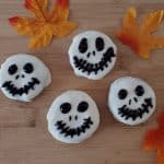 Four Halloween Chocolate Covered Oreos decorated with white icing and black icing Jack Skellington faces rest on a wooden surface, surrounded by three artificial autumn leaves.