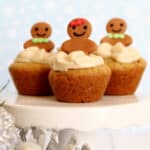 Three Gingerbread Cookie Cups topped with gingerbread person decorations and swirls of frosting, displayed on a white cake stand against a soft blue, festive background. Sparkling silver decorations are visible in the foreground.