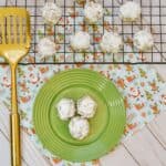 Three powdered sugar-coated cookies on a green plate, with more cookies cooling on a wire rack nearby. A gold spatula and a festive napkin with holiday designs add to the scene—perfect for christmas dessert ideas on a light wooden surface.
