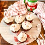 Six hot chocolate cupcakes with white frosting, mini marshmallows, and striped straws sit on a white round board. A festive snowman mug and red-and-white checkered towels are in the background on a wooden surface.