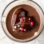 A glass mug filled with hot chocolate, topped with a heart-shaped chocolate piece decorated with small red and white sprinkles—this cozy valentines hot chocolate bombs treat sits on a white tiled surface.