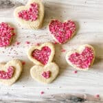 Heart-shaped sugar cookies topped with pink, red, and white sprinkles are arranged on a light wooden surface. Some cookies have a heart outline filled with sprinkles, while others are fully covered.