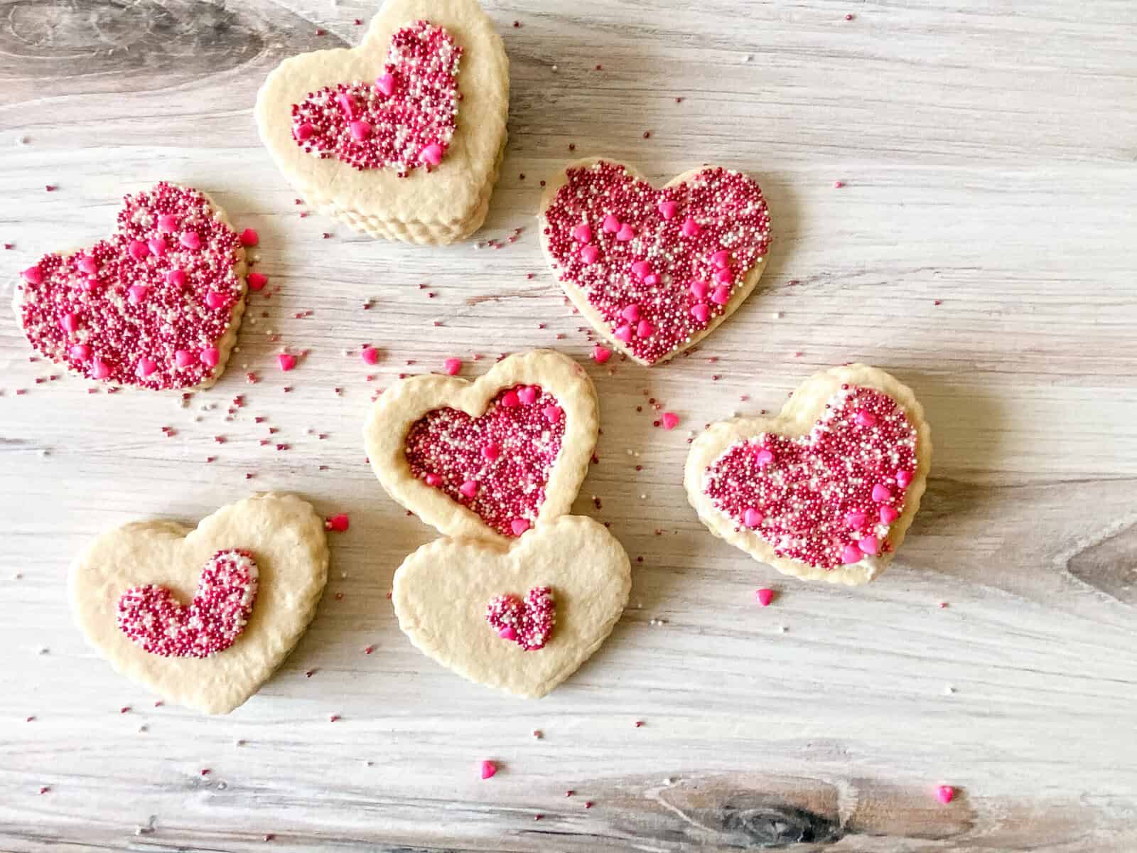 Heart-shaped sugar cookies topped with pink, red, and white sprinkles are arranged on a light wooden surface. Some cookies have a heart outline filled with sprinkles, while others are fully covered.