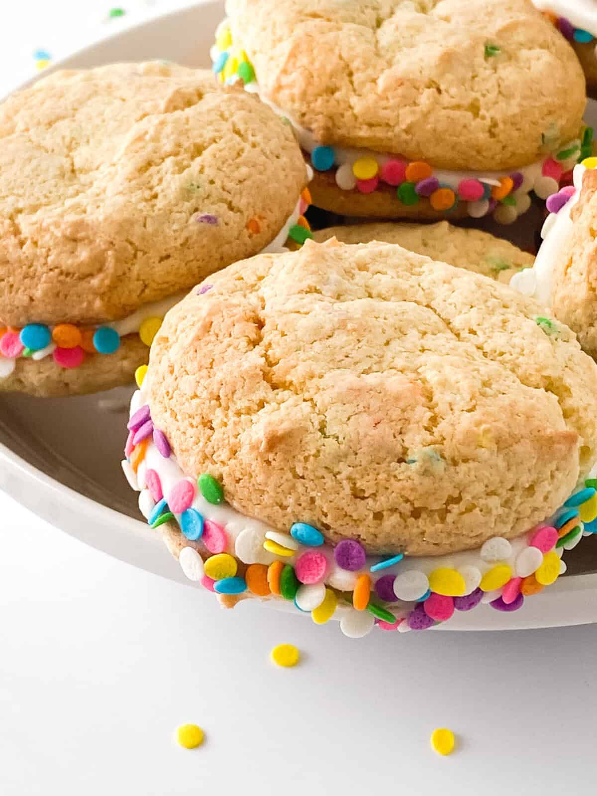 Three ice cream sandwiches made with funfetti cookies—perfect for recipes using yellow cake mix—and rolled in colorful round sprinkles are stacked on a plate, with extra sprinkles scattered nearby.