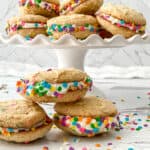 Whoopie pies with white frosting and colorful confetti sprinkles are stacked on a white cake stand, showcasing one of our favorite whoopie pie recipes, with more pies and scattered sprinkles on a marble surface in the foreground.