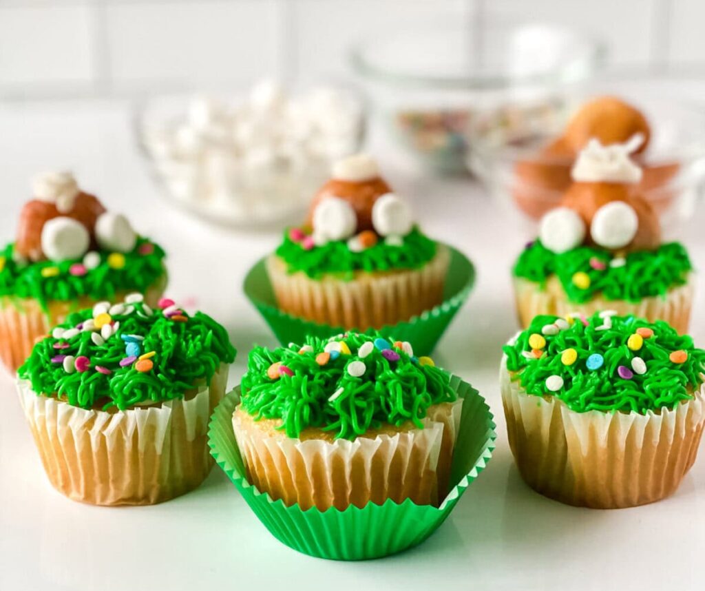 Six cupcakes with green frosting resembling grass, decorated with colorful sprinkles and marshmallows, are displayed on a white surface. Baking ingredients are blurred in the background.