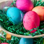 A close-up of a white dish filled with green plastic Easter grass and five brightly colored Easter eggs, showcasing vibrant pink, blue, orange, and purple hues—perfect inspiration for learning how to dye eggs with food coloring.