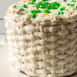 A round cake decorated with elegant basket cake design in white frosting, topped with white swirls, green sprinkles, and small, colorful round sprinkles, displayed on a white scalloped plate.