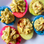 A plate of colored deviled eggs with pink, blue, and yellow dyed egg whites, filled with a creamy yellow yolk mixture and sprinkled with paprika.