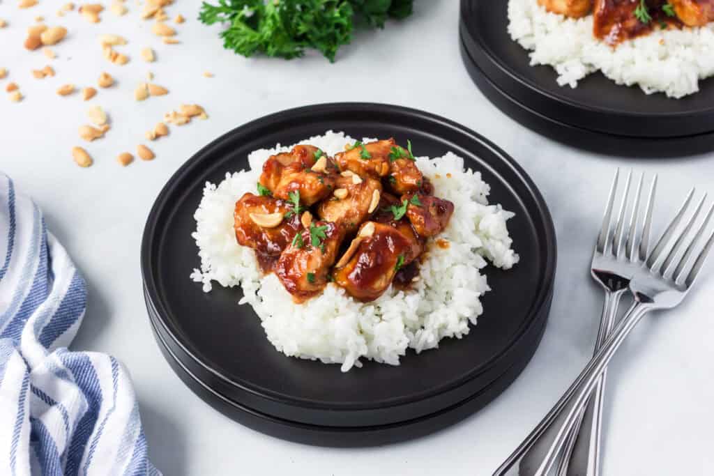 A black plate with white rice topped with saucy peanut butter chicken and chopped peanuts, garnished with herbs. Another similar plate, forks, and a blue striped napkin are nearby on the table.