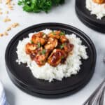 A black plate with white rice topped with saucy peanut butter chicken and chopped peanuts, garnished with herbs. Another similar plate, forks, and a blue striped napkin are nearby on the table.