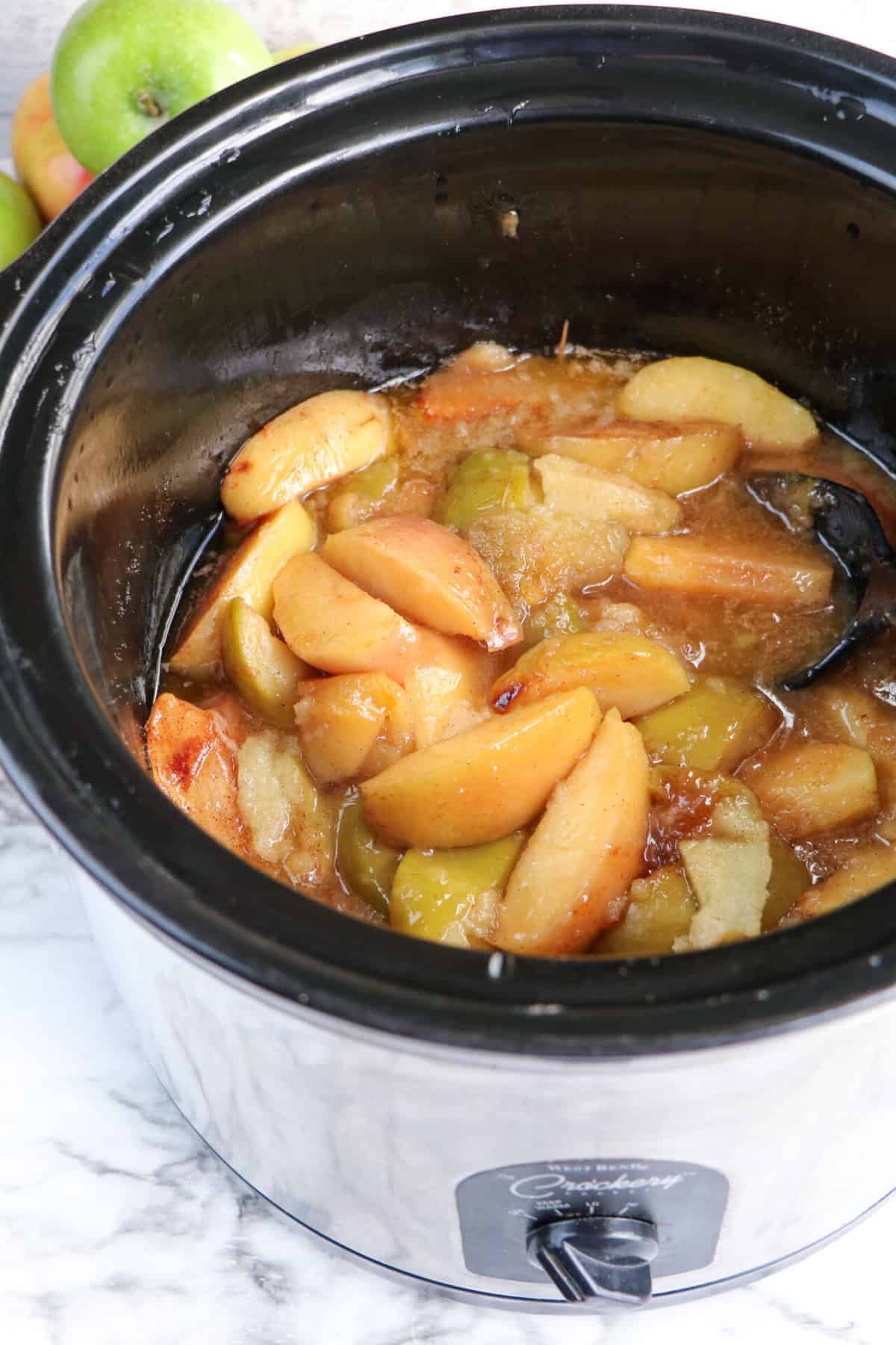 Slices of cooked apples in a sweet sauce inside a black slow cooker pot, with some green apples visible in the background on a marble surface.