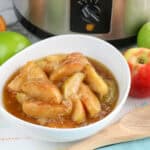 A bowl of cooked cinnamon apples in syrup sits on a striped cloth, surrounded by fresh apples, with a wooden spoon and slow cooker in the background—perfect for festive 4th of July Crockpot Meals.