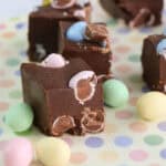 Squares of chocolate fudge topped with pastel-colored candy eggs sit on a pastel polka dot surface, with more candy eggs scattered around. This Easter Fudge Recipe looks rich and creamy, with crisp eggs adding festive texture.