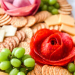 Charcuterie board with crackers, cheese, grapes, and salami and ham arranged like roses.