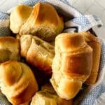 A basket lined with a blue and white towel holds several golden-brown, fluffy homemade crescent rolls on a white tiled surface.