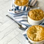 Three Mini Chicken Pot Pies in white ramekins, with golden brown crusts, are arranged on a blue and white striped cloth alongside a silver spoon on a light marble surface.