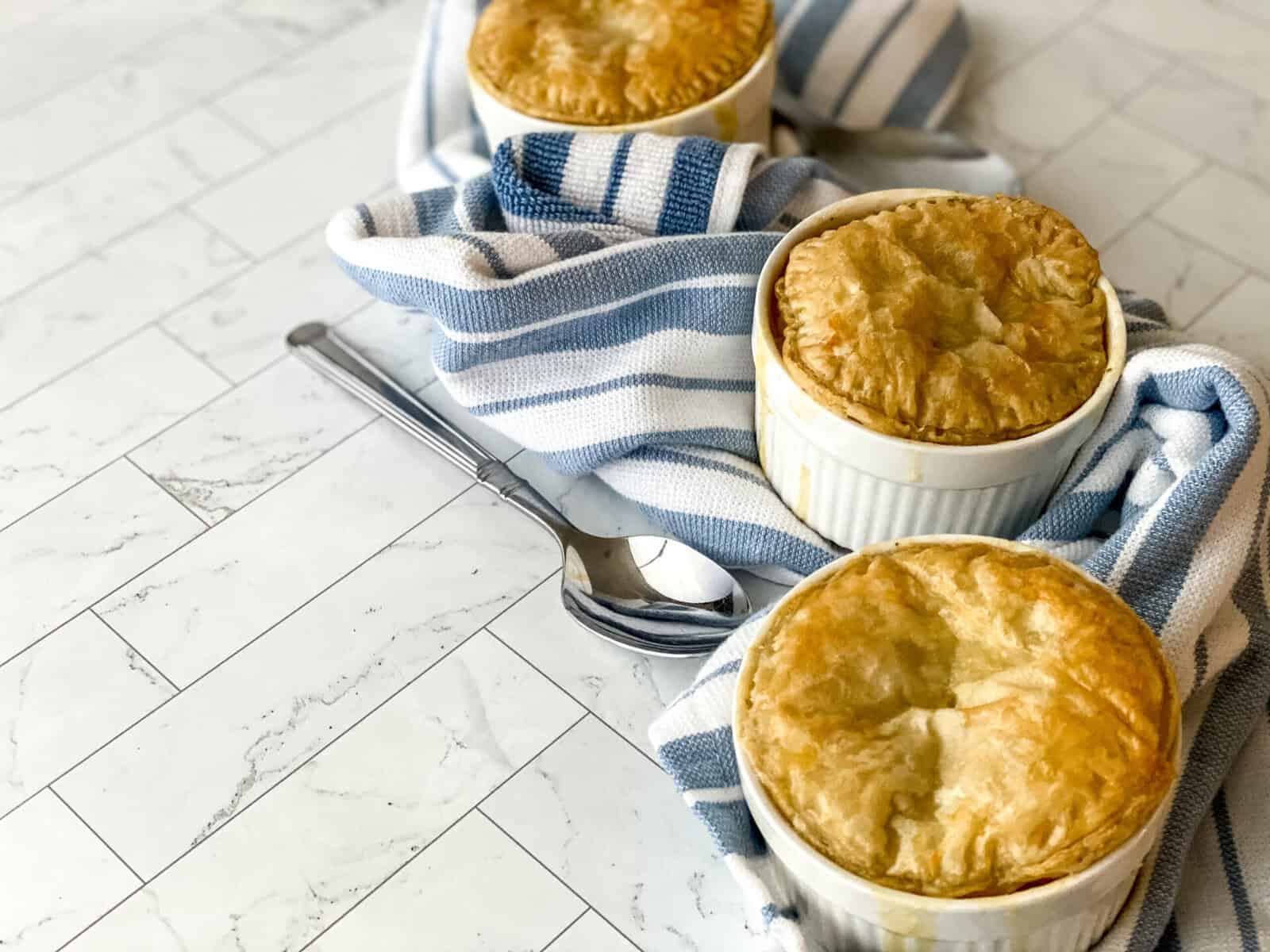 Three Mini Chicken Pot Pies in white ramekins, with golden brown crusts, are arranged on a blue and white striped cloth alongside a silver spoon on a light marble surface.