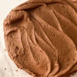 A close-up view of a round chocolate cake with creamy chocolate buttercream frosting recipe, swirled smoothly on top, sitting on a white surface.