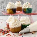 Five cupcakes with colorful wrappers and white frosting are displayed on pink stands and a marble surface, surrounded by rainbow sprinkles and a piping bag filled with frosting.