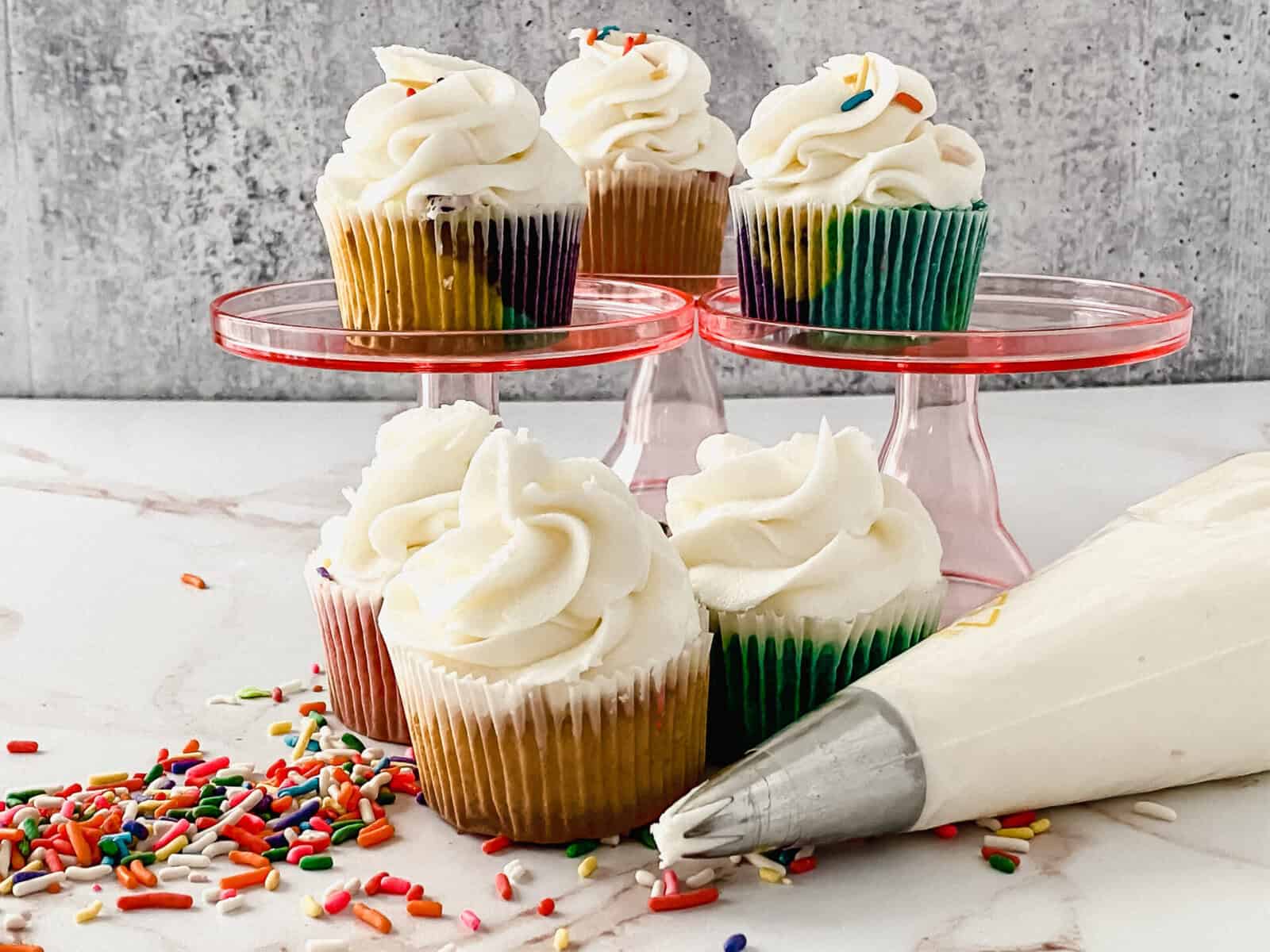 Five cupcakes with colorful wrappers and white frosting are displayed on pink stands and a marble surface, surrounded by rainbow sprinkles and a piping bag filled with frosting.