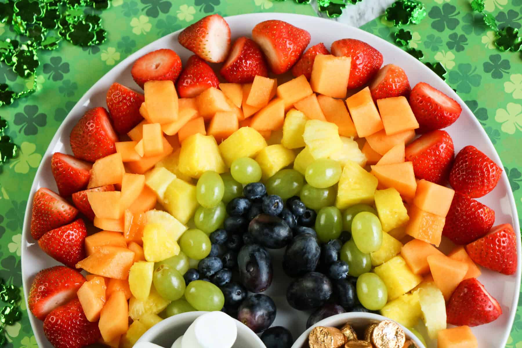 A plate arranged with strawberries, cantaloupe, pineapple, green grapes, and blueberries forms a vibrant fruit rainbow, surrounded by green clover decorations on a festive table.