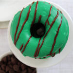 A green-frosted donut with chocolate drizzle sits on a white plate, viewed from above—a festive choice for St Patrick’s Day Desserts. In the background, there’s a bowl of chocolate chips and part of another plate.