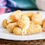 A white plate with golden-brown breaded mozzarella sticks, some with melted cheese oozing out, sits on a woven placemat—perfect Easy Appetizers for a Party—with a colorful cloth in the background.
