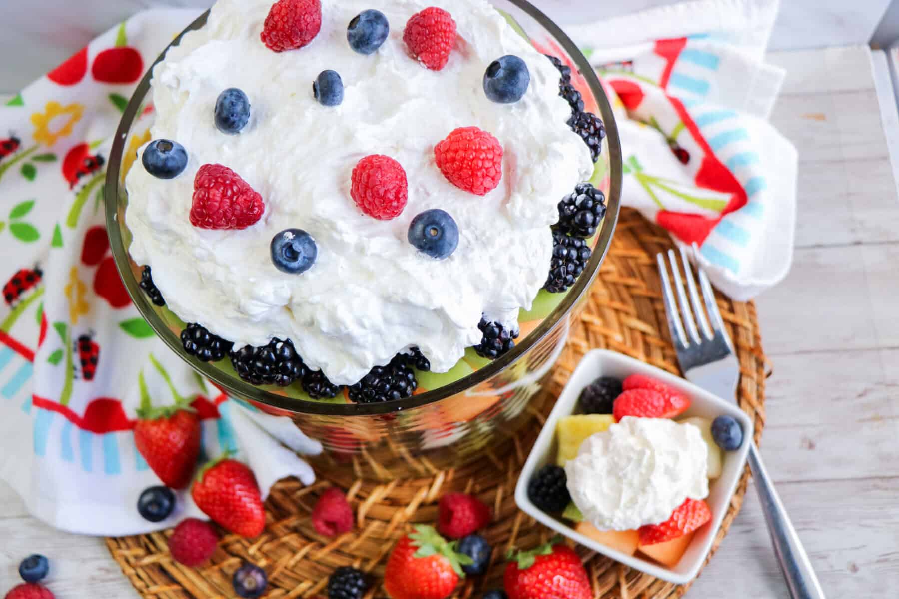 A large glass trifle bowl filled with layers of fruit, topped with cool whip fruit dip and fresh berries. Nearby, a small dish holds a serving of the dessert, surrounded by scattered strawberries and blueberries.