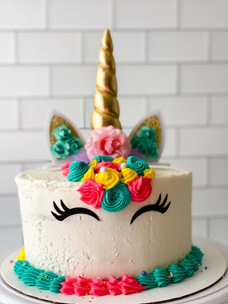A round white cake decorated as a unicorn with a golden horn, ears, closed eyes with long lashes, and colorful icing flowers in pink, yellow, and teal on top. The background is white tile.