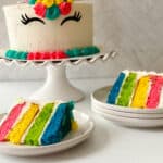 A rainbow unicorn cake with colorful frosting and closed-eye designs sits on a stand. Two plates hold slices revealing bright pink, yellow, green, and blue layers wrapped in white icing.