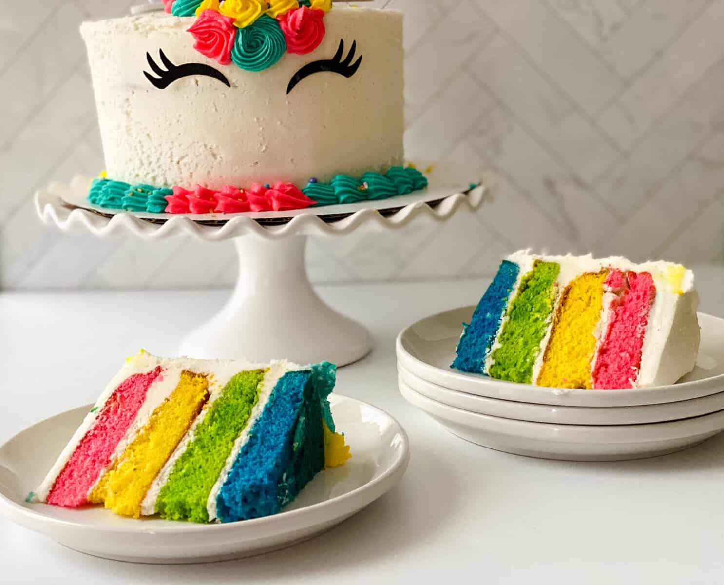 A rainbow unicorn cake with colorful frosting and closed-eye designs sits on a stand. Two plates hold slices revealing bright pink, yellow, green, and blue layers wrapped in white icing.