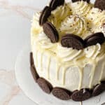 A round cake with white frosting and white chocolate drip, topped with swirls of cream, chocolate sandwich cookies, and sprinkles. More cookies line the sides—perfect for anyone searching for unique birthday cakes ideas.