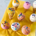 Ice cream cone cake pops are arranged upright on a yellow napkin, coated in white, pink, and purple chocolate, and finished with colorful sprinkles and drizzles.