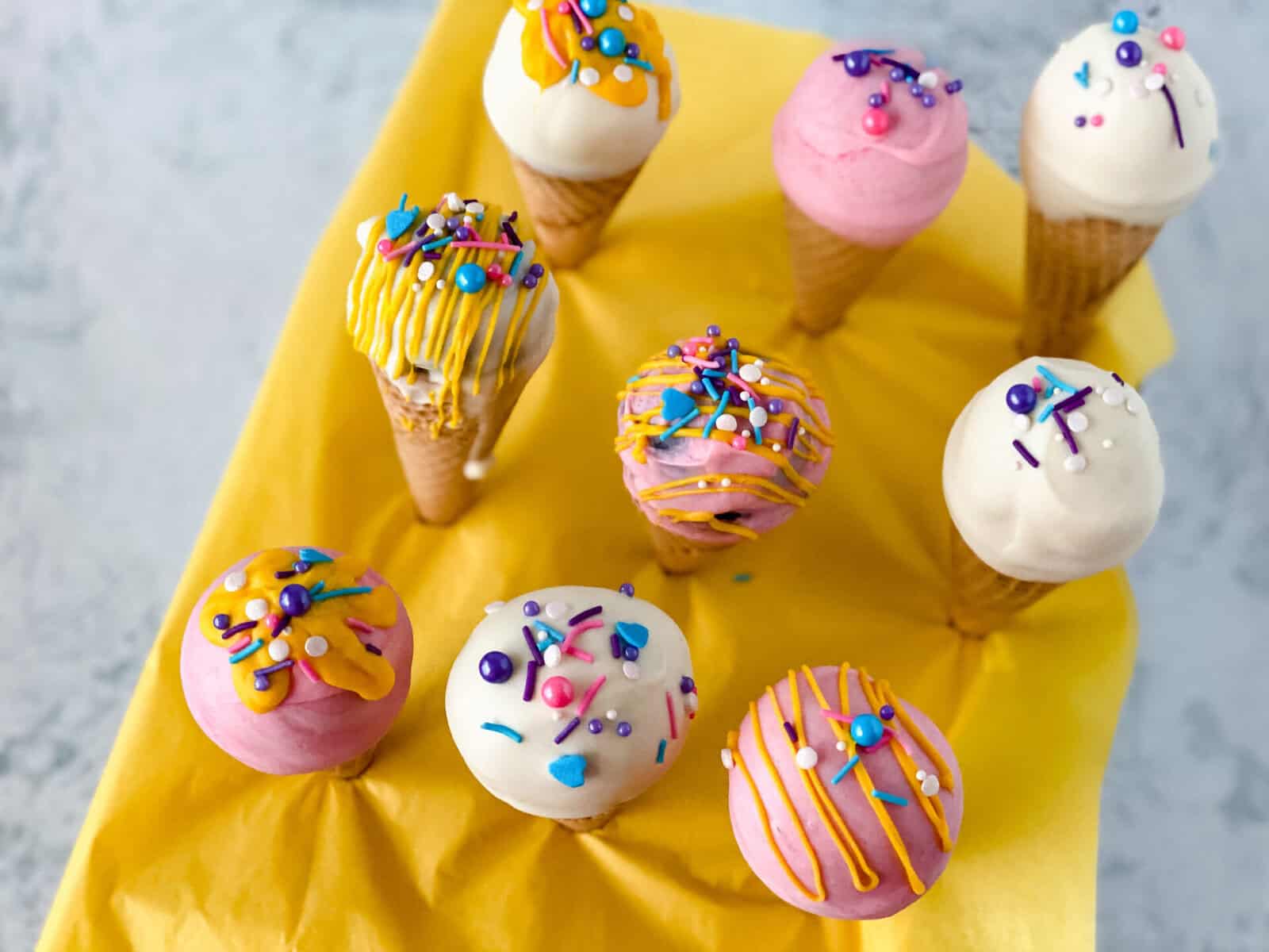 Ice cream cone cake pops are arranged upright on a yellow napkin, coated in white, pink, and purple chocolate, and finished with colorful sprinkles and drizzles.