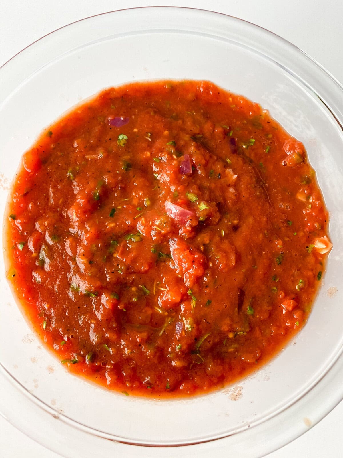 A glass bowl filled with chunky red salsa made from a fresh salsa recipe, showcasing visible pieces of tomato, onion, and green herbs, placed on a white surface.