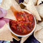 A hand holds a triangular tortilla chip dipped in chunky red salsa, surrounded by more tortilla chips and a small bowl of salsa—deliciously simple, these are perfect easy appetizers for a party.