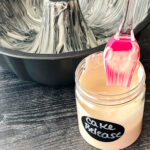 A pink brush dipped in a jar of cake release next to a greased bundt pan.
