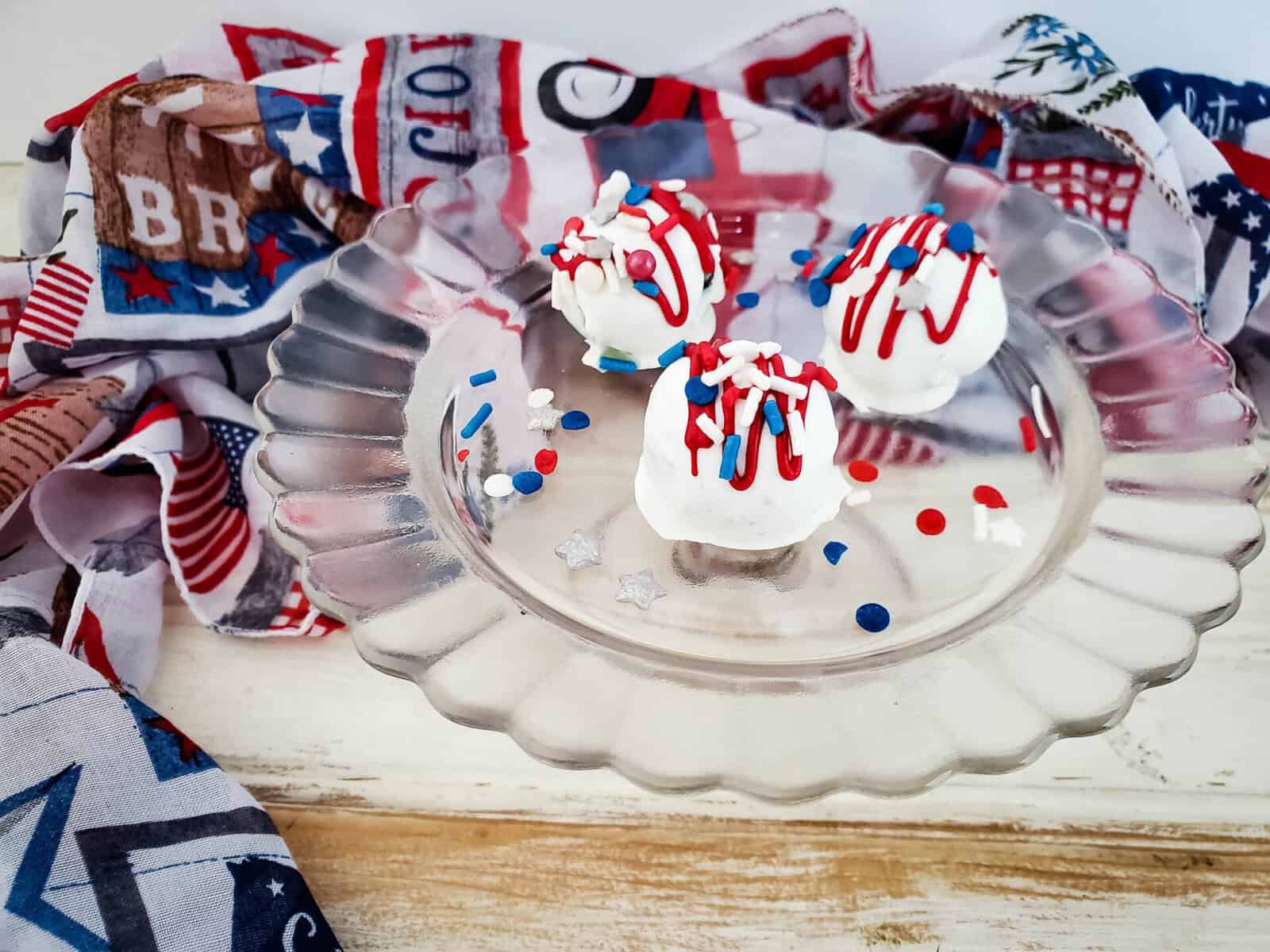 Three Red White Blue Oreo Balls, decorated with festive sprinkles and drizzle, sit on a glass plate. Patriotic-themed fabric with stars and stripes adds a celebratory touch in the background.