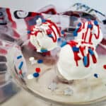 Three Red White Blue Oreo Balls, coated in white chocolate and topped with festive red, white, and blue sprinkles and drizzle, sit on a clear glass plate. Extra sprinkles are scattered around, with a patriotic-themed cloth in the background.