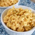 A white bowl filled with creamy macaroni and cheese, inspired by the best mac and cheese recipes, is garnished with herbs and sits on a blue and white patterned surface. Another similar bowl is partially visible in the background.