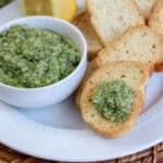 A white plate holds slices of toasted bread topped with easy homemade pesto. A small white bowl filled with pesto and a sliced lemon rest on the plate, set on a woven placemat.