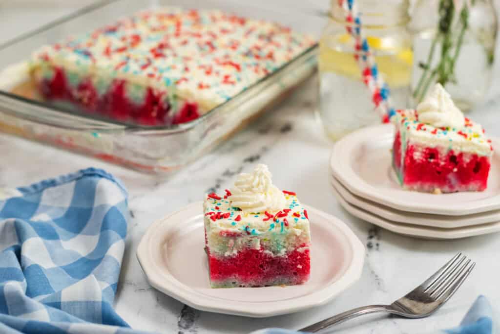A slice of red white and blue poke cake topped with whipped cream and sprinkles sits on a plate, with the rest of the festive cake in a glass dish behind it. A blue checkered napkin and a glass of lemonade are nearby.